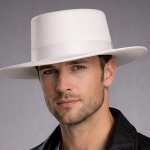Op de afbeelding: Een witte fedora hoed met een platte bovenkant en een brede rand. De hoed heeft een witte band rond de kroon. Deze klassieke hoed wordt vaak gedragen voor formele gelegenheden of om een vleugje stijl aan een outfit toe te voegen.