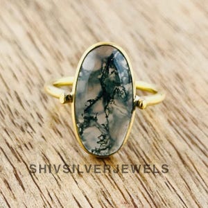 Puede incluir: Anillo dorado con una piedra ovalada de ágata musgosa, con patrones verdes y blancos. El anillo está sobre una superficie de madera. El texto "SHIVSILVERJEWELS" es visible.