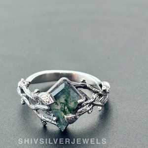 Puede incluir: Un anillo de plata con una piedra preciosa de ágata verde musgo engastada en un diseño de ramas. El anillo está sobre un fondo gris. SHIVSILVERJEWELS está impreso debajo del anillo.
