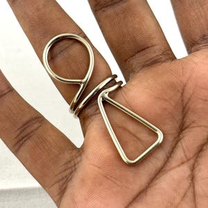 May include: A silver-toned ring with a unique geometric design. The ring features a circular loop, a triangular shape, and multiple bands. The ring is worn on a finger, showcasing its modern and artistic style.
