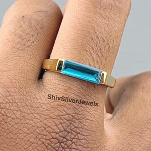 Könnte beinhalten: Ein goldener Ring mit einem blauen rechteckigen Edelstein. Der Ring wird an einem Finger getragen.