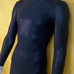 Op de afbeelding: Zwarte bodysuit met lange mouwen en een textuur met honingraatpatroon. Het kledingstuk heeft een ronde hals en een nauwsluitend ontwerp. De stof heeft een matte afwerking en de bodysuit wordt op een mannequin gepresenteerd tegen een gele achtergrond.
