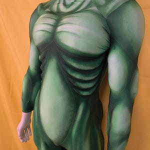 Può includere: Un body verde con un design muscolare, con ombreggiatura per creare l'illusione di un fisico muscoloso. Il costume ha un collo alto e maniche lunghe, con dettagli che imitano l'aspetto di un busto e degli arti.
