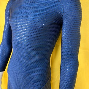 Puede incluir: Un body azul con un diseño texturizado y estampado. La prenda de manga larga tiene un cuello redondo y se muestra en un maniquí sobre un fondo amarillo. La tela parece tener un brillo sutil.