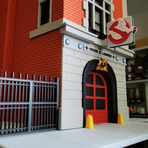 Op de afbeelding: Een miniatuurreplica van het Ghostbusters-brandweerkazerne, met een rode bakstenen gevel, witte afwerking en een zwarte poort. Het iconische Ghostbusters-logo staat op een bord. Het gebouw heeft een rode deur en de tekst "-6 HOOK & LADDER-6-".