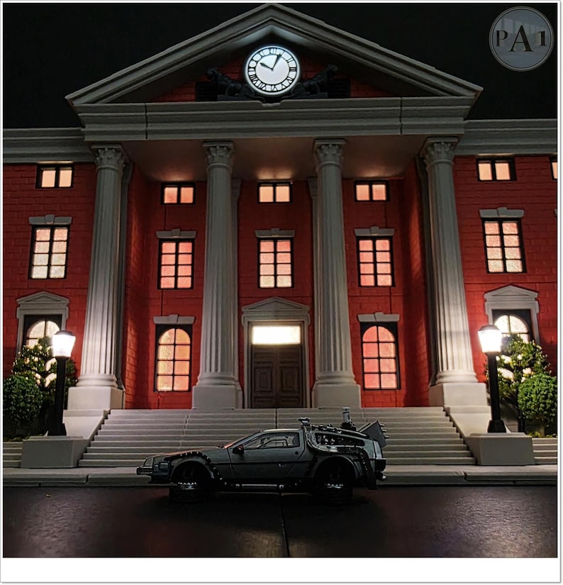 May include: A miniature model of a red brick building with white columns and a clock, illuminated by warm lighting. A silver DeLorean car is parked in front of the building, creating a scene reminiscent of a movie.
