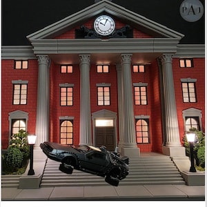 May include: A detailed model of a red brick building with white columns and a clock tower. A black DeLorean car is suspended in mid-air in front of the building. The clock shows the time. The scene is lit with warm interior lights.