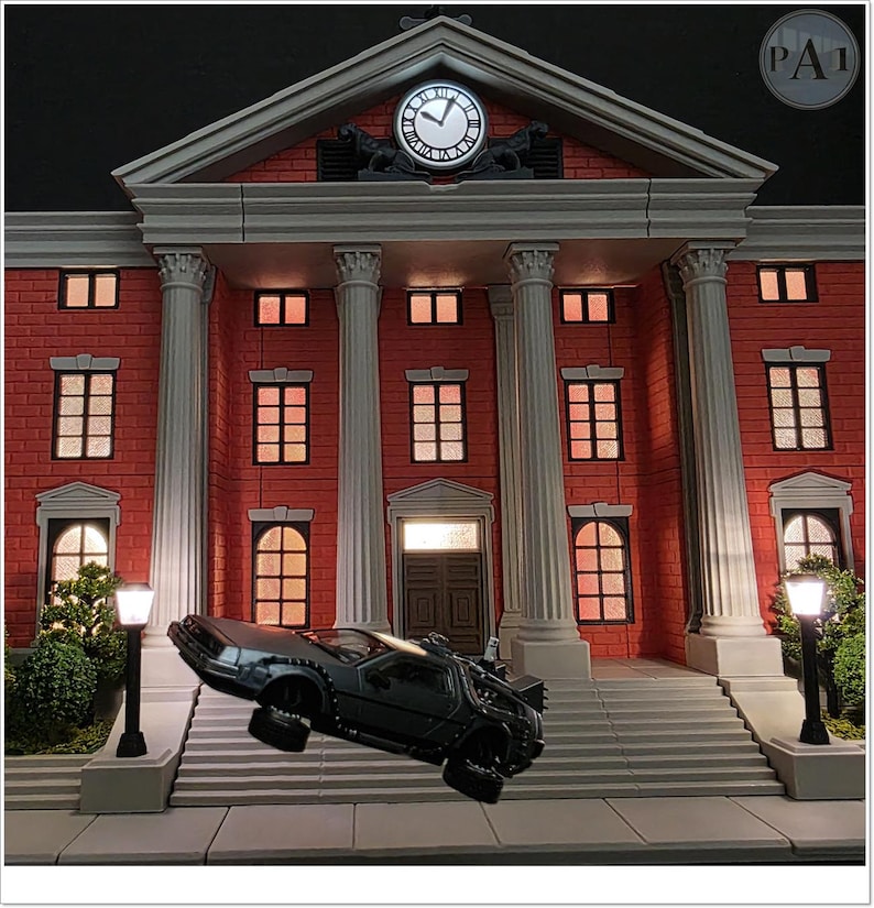 May include: A detailed model of a red brick building with white columns and a clock tower. A black DeLorean car is suspended in mid-air in front of the building. The clock shows the time. The scene is lit with warm interior lights.