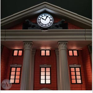 May include: A detailed model of a building facade, featuring a clock tower with a glowing clock face. The building has a red brick exterior, white columns, and illuminated windows. Two statues are on the roof. The clock shows the time.