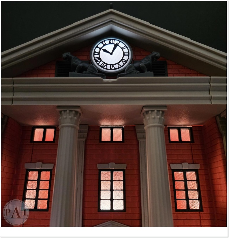 May include: A detailed model of a building facade, featuring a clock tower with a glowing clock face. The building has a red brick exterior, white columns, and illuminated windows. Two statues are on the roof. The clock shows the time.