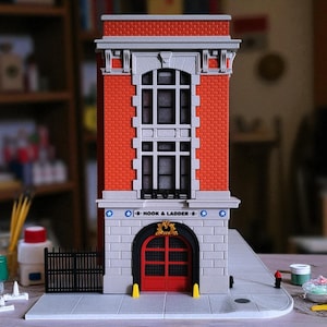Può includere: Un edificio in miniatura in mattoni rossi con una facciata in pietra grigia e una porta rossa. L'edificio ha un'insegna con la scritta "Hook & Ladder 8". C'è una recinzione nera e un marciapiede.