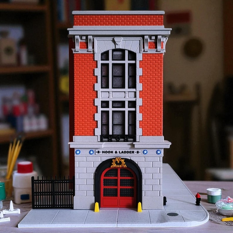 Op de afbeelding: Een miniatuurgebouw van rode baksteen met een grijze stenen gevel en een rode deur. Het gebouw heeft een bord met de tekst "Hook & Ladder 8". Het heeft een zwart hek en een stoep.