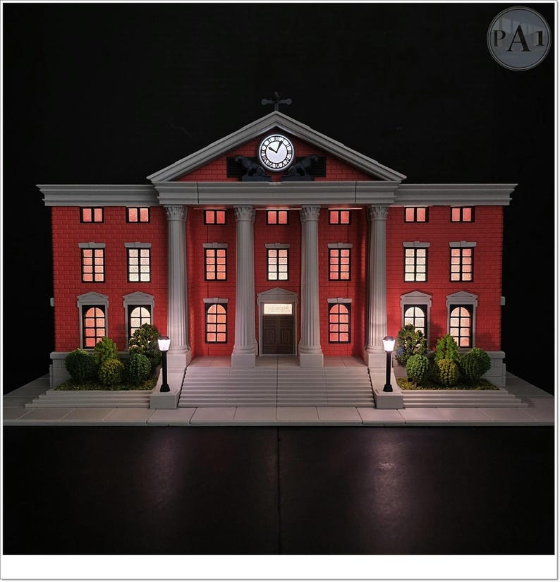 May include: A miniature replica of a red brick building with white columns and a clock tower. The building has multiple windows with interior lighting, and small bushes and street lamps are in front. The clock shows the time.