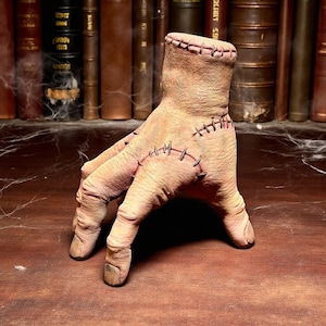 Puede incluir: Una mano de utilería con un diseño cosido, con una muñeca cortada y puntadas visibles. La mano está colocada sobre una superficie de madera, con un telón de fondo de libros antiguos y un toque de telarañas, creando una estética espeluznante con temática de Halloween.