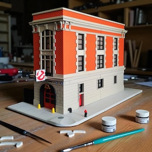 Op de afbeelding: Een gedetailleerd model van het Ghostbusters-brandweerhuis, met rode bakstenen muren, witte afwerking en het Ghostbusters-logo. Het model bevat een rode deur, ramen en een stoepbasis. Penselen en verfpotten zijn in de buurt.