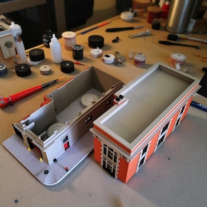 Op de afbeelding: Twee miniatuur gebouwmodellen, waarvan &eacute;&eacute;n open om interieurdetails te onthullen. De gebouwen hebben oranje bakstenen gevels, witte randen en grijze daken. Diverse gereedschappen en verfbenodigdheden omringen de modellen op een houten oppervlak.