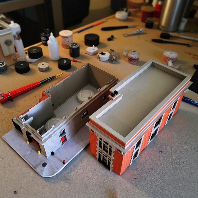 Op de afbeelding: Twee miniatuur gebouwmodellen, waarvan &eacute;&eacute;n open om interieurdetails te onthullen. De gebouwen hebben oranje bakstenen gevels, witte randen en grijze daken. Diverse gereedschappen en verfbenodigdheden omringen de modellen op een houten oppervlak.