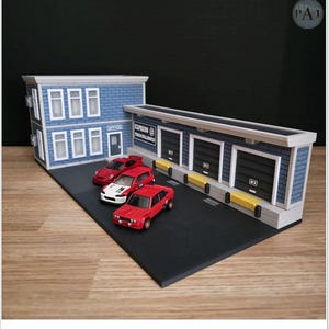 Puede incluir: Un diorama en miniatura detallado con un edificio de dos pisos y un almacén con tres puertas de garaje. Tres pequeños coches rojos están aparcados sobre una superficie negra. El almacén tiene el texto "EXPRESS TRUCKLINES".