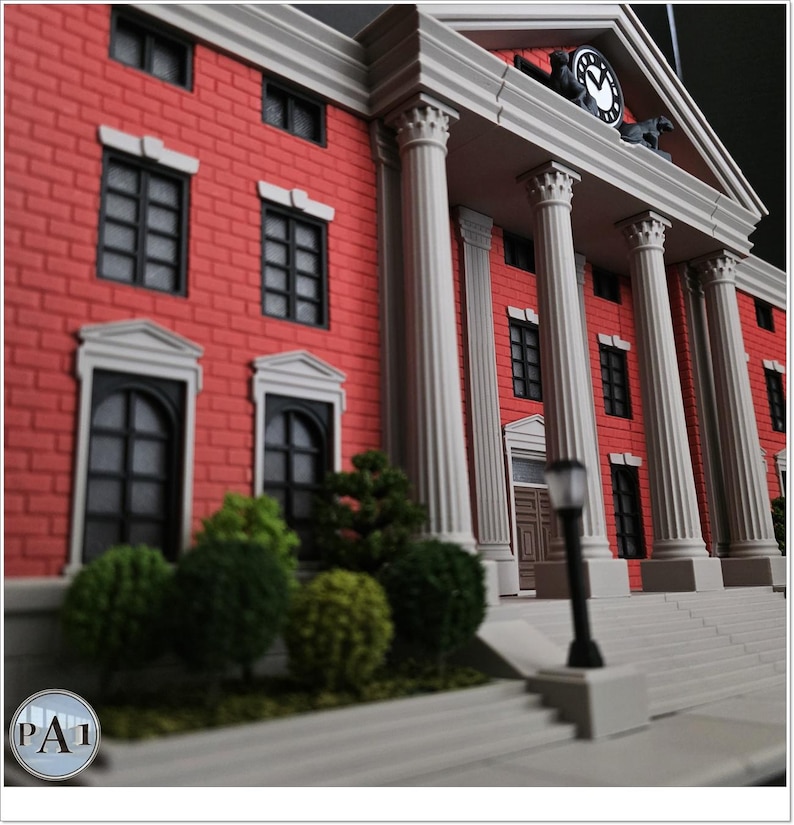 May include: A miniature model of a red brick building with white columns and a clock tower. The building has black windows and doors, with green shrubbery in front. The clock tower has a clock face and two horse figures.