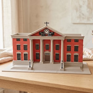 May include: A detailed miniature model of a brick building with white columns, black windows, and a clock tower. The model is red and grey, with a cross on top. It sits on a light grey base, with two small street lamps.