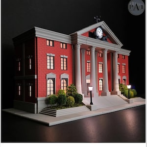 May include: A miniature model of a red brick building with white columns and a clock tower. The model has lit windows and street lamps, and a small green bush at the base of the building. The model is a replica of a courthouse.