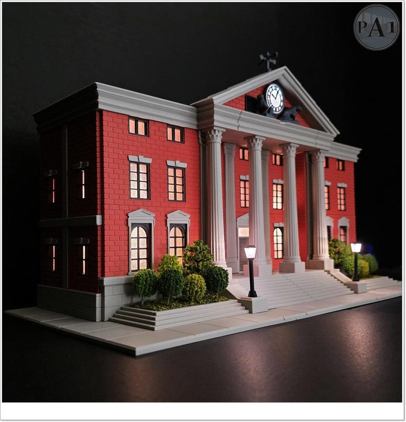 May include: A miniature model of a red brick building with white columns and a clock tower. The model has lit windows and street lamps, and a small green bush at the base of the building. The model is a replica of a courthouse.