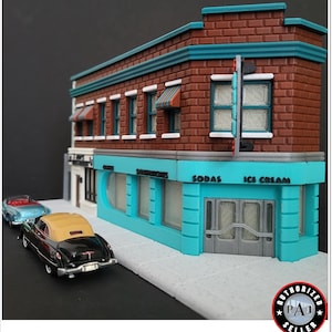 May include: A miniature model of a 1950s-style ice cream shop with a turquoise facade, red brick exterior, and white trim. The shop has a sign that reads "Ice Cream, Sodas, Sandwiches". There are two vintage cars parked in front of the shop.