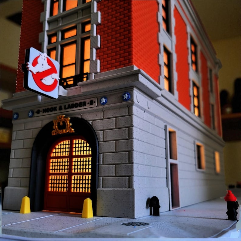 Op de afbeelding: Gedetailleerd model van een bakstenen gebouw met een bord met een spooklogo. Het gebouw heeft een grijze stenen gevel, rode bakstenen bovenverdiepingen en verlichte ramen. Het bord luidt "HOOK & LADDER 8".