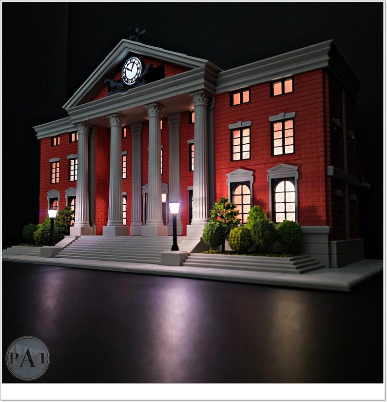 May include: A miniature model of a red brick building with white columns, a clock, and illuminated windows. The building has a classical design with a pediment and a clock tower. Small trees and street lamps are in front of the building.