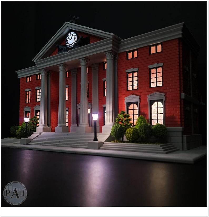 May include: A miniature model of a red brick building with white columns and a clock tower. The building has multiple windows with interior lighting, and is flanked by small trees and street lamps. The model is set against a dark background.