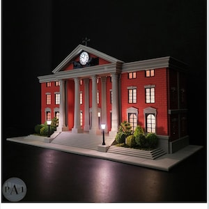 May include: A miniature model of a red brick building with white columns, a clock, and illuminated windows. The model features a detailed facade, a cross on top, and small street lamps. The overall design is reminiscent of a courthouse.