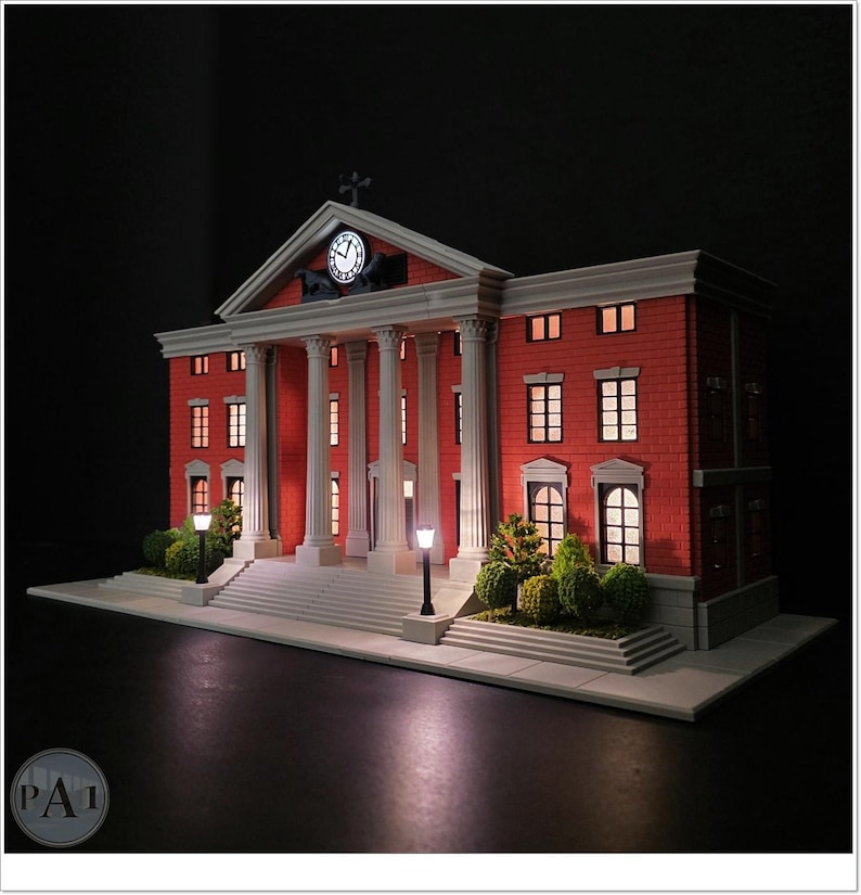 May include: A miniature model of a red brick building with white columns, a clock, and illuminated windows. The model features a detailed facade, a cross on top, and small street lamps. The overall design is reminiscent of a courthouse.