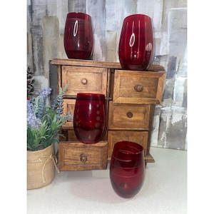 May include: Four stemless red glass tumblers with etched patterns, including horizontal lines and circular motifs, arranged around a small wooden chest with multiple drawers and a potted lavender plant in a burlap-wrapped container.