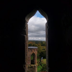 May include: A view from a stone window looking out at a stone tower with a rounded roof and a small arched opening. The tower is surrounded by green trees and a small stream. The sky is cloudy and blue.