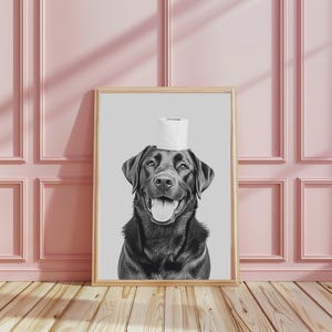 May include: A framed black and white art print featuring a portrait of a black Labrador retriever with a roll of white toilet paper balanced on its head. The dog has a happy expression with its mouth open and tongue visible against a gray background.