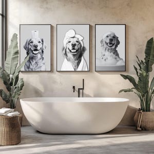 May include: A set of three black and white framed wall art prints featuring golden retriever dogs. The first shows a dog with a toilet paper roll on its head, the second a dog in a bathrobe and towel wrap, and the third a dog in a bubble bath.