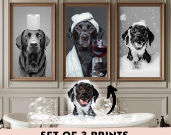 May include: Set of three framed prints featuring black Labrador Retrievers in humorous poses. One dog has a toilet paper roll on its head, another holds a wine glass, and the third is in a bubble bath. The prints are displayed in a bathroom setting.