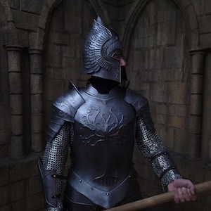 Puede incluir: Un caballero con armadura completa, que lleva un casco con alas, sostiene un bastón de madera. La armadura es plateada y tiene diseños intrincados. El caballero está de pie frente a una pared de piedra.