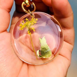 Op de afbeelding: Een heldere, ronde sleutelhanger met een goudkleurige hartvormige sluiting. Binnenin de heldere hars bevindt zich een gele bloem en een groen katten silhouet. De sleutelhanger wordt in een hand gehouden.