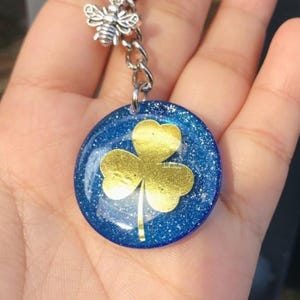 Op de afbeelding: Een sleutelhanger met een ronde, blauwe hanger met een gouden klaverontwerp. De hanger is bevestigd aan een zilveren ketting en een bijenbedel. De sleutelhanger wordt vastgehouden door een zilveren sleutelring.