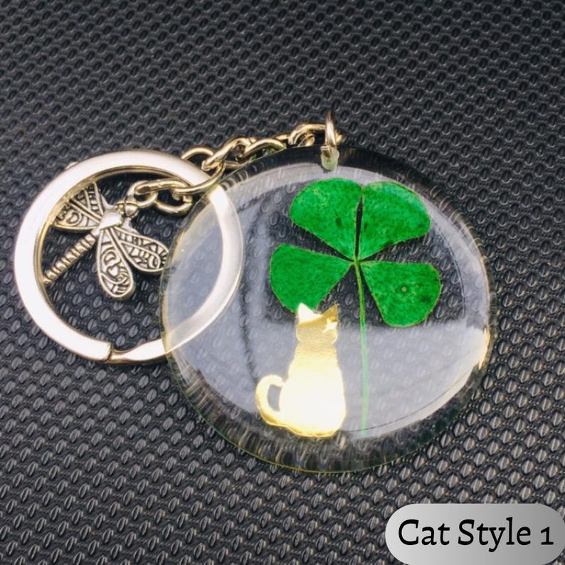 Op de afbeelding: Een heldere, ronde sleutelhanger met een goudkleurig katten silhouet en een groene klaver met vier bladeren. De sleutelhanger heeft een zilveren libellenbedel en de tekst "Cat Style 1".