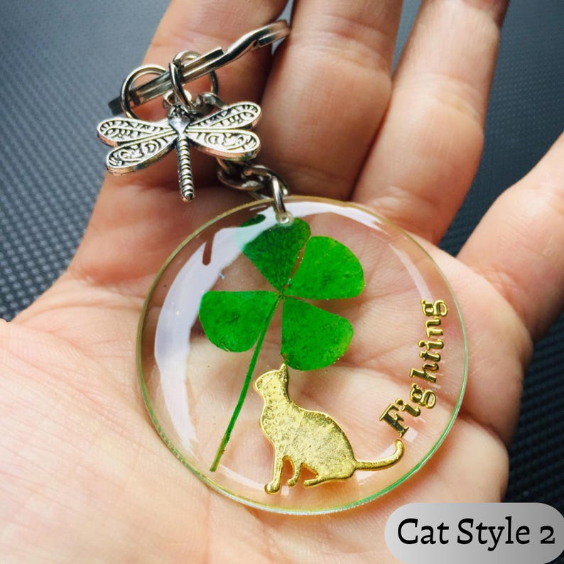 Op de afbeelding: Een heldere hars sleutelhanger met een klavertjevier en een gouden kattenbedel. De sleutelhanger heeft het woord "Fighting" in gouden letters en een zilveren libellenbedel. De tekst "Cat Style 2" staat onderaan.