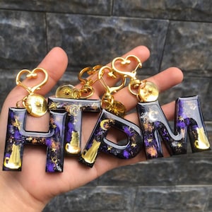 Op de afbeelding: Sleutelhangers met de letters H, T, P en M. Elke letter is een andere sleutelhanger met een goudkleurige metalen sluiting en een goudkleurige letter. De letters zijn gevuld met een paarse en zwarte hars met gouden en witte accenten. De sluitingen hebben hart- en zonontwerpen.