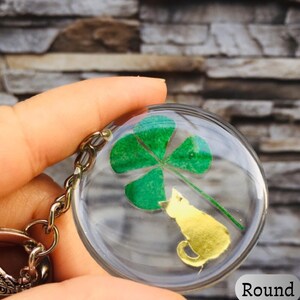 Op de afbeelding: Ronde sleutelhanger met een doorzichtige harskoepel. Binnenin de koepel zit een groene klaver met vier bladeren en een goudkleurig katten silhouet. De sleutelhanger heeft een zilverkleurige ketting en sluiting.