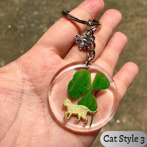 Op de afbeelding: Transparante hars sleutelhanger met een klavertjevier en een goudkleurige kattenbedel. De sleutelhanger heeft een zilverkleurige bijenbedel en een metalen ring. De tekst "Cat Style 3" staat rechtsonder.
