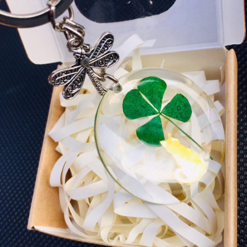 Op de afbeelding: Een heldere hars sleutelhanger met een klavertjevier en een kleine gele bloem. De sleutelhanger heeft een zilveren libellenbedel en wordt gepresenteerd in een kleine kartonnen doos met witte papieren vulling.