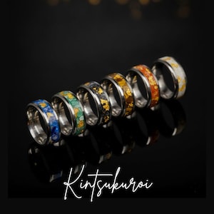 May include: A collection of six metal rings arranged in a diagonal row, each featuring a central inlay of crushed stone and gold-colored flakes. The rings display various colors including blue, green, black, brown, orange, and white. The text Kintsukuroi is visible.