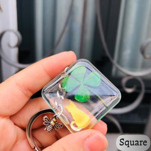Op de afbeelding: Vierkante sleutelhanger met een heldere harsbehuizing. Binnenin een groene klaver met vier bladeren en een gele kat-silhouet. Een zilverkleurige bijenbedel en een sleutelring zijn bevestigd. Het woord "Square" is wit gedrukt.
