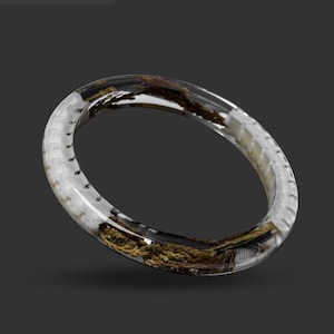 Pode incluir: Uma pulseira de resina transparente com elementos naturais embutidos. A pulseira apresenta uma mistura de materiais orgânicos brancos, dourados e castanhos, criando uma aparência única e texturizada. O design é circular e tem um acabamento liso e polido.