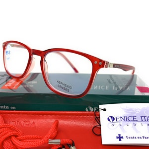 May include: Red plastic framed reading glasses with aspheric lenses. The glasses are on top of a green and white box with the text "Venice Italy" and "occhiali" on a white tag. The box also has the text "Venta en Farmacias".